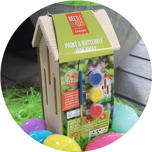 A a wooden butterfly habitat with a set of paints and brushes attached, all surrounded by green easter grass and colorful easter eggs.